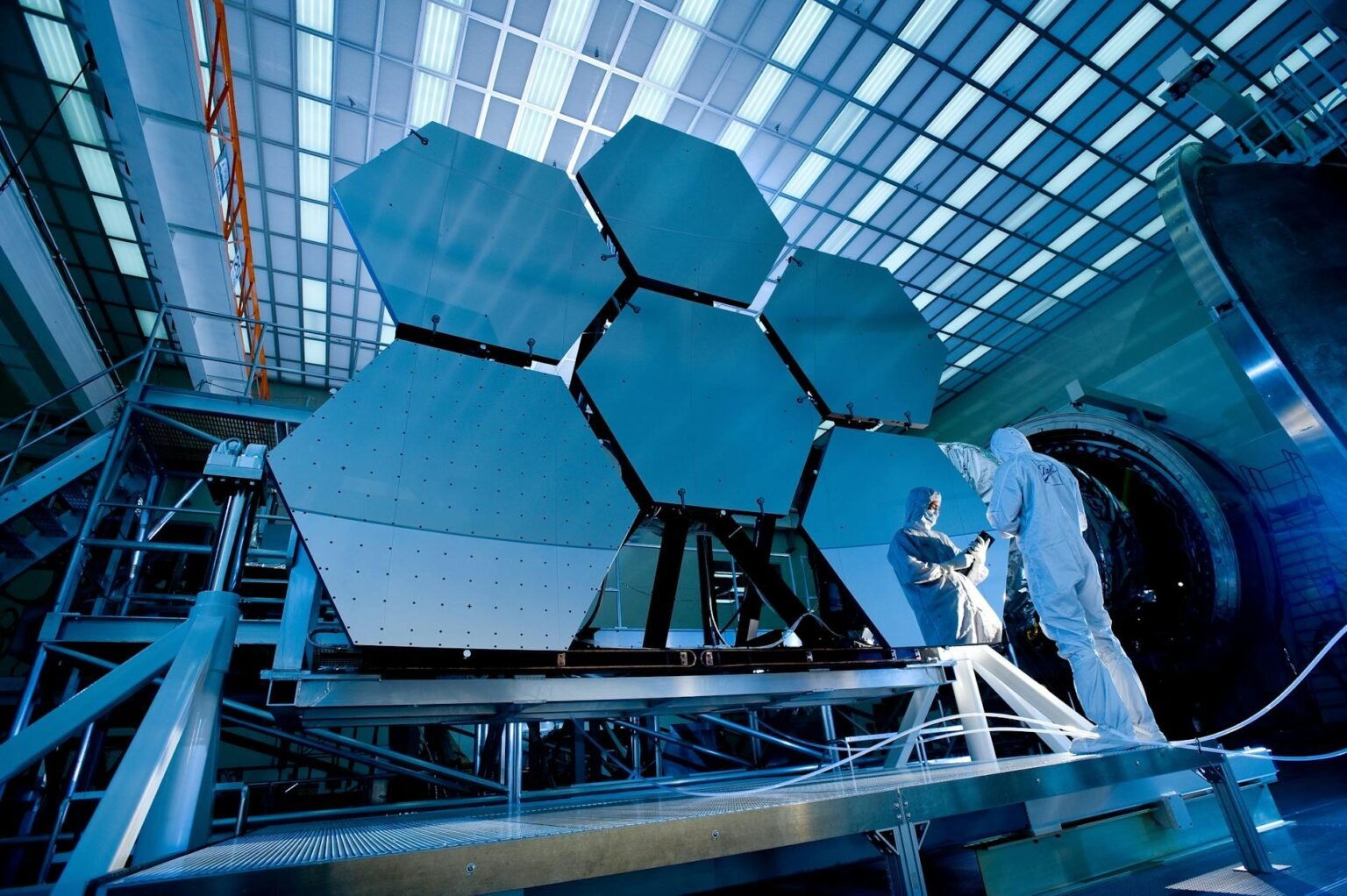 Engineers in protective suits work on telescopic mirrors in a high-tech lab.